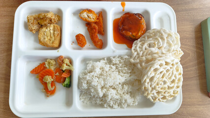 A white compartmentalized lunch tray filled with various Indonesian-style dishes including rice, fried chicken, vegetables, a sauced patty, and krupuk crackers on a wooden table.