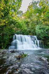 Fototapeta premium Lush green forest canopy framing beautiful cascading waterfall with sun starburst shining through dense foliage, creating tranquil natural landscape scene