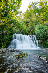 Fototapeta premium Serene tropical waterfall cascading in silky long exposure through lush green foliage, sun filtering through the canopy creating warm rays over clear flowing water and mossy rocks