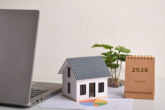 Miniature house with laptop and 2026 desk calendar on workspace. Real estate planning, property investment strategy, housing market outlook, and home buying financial management concept.