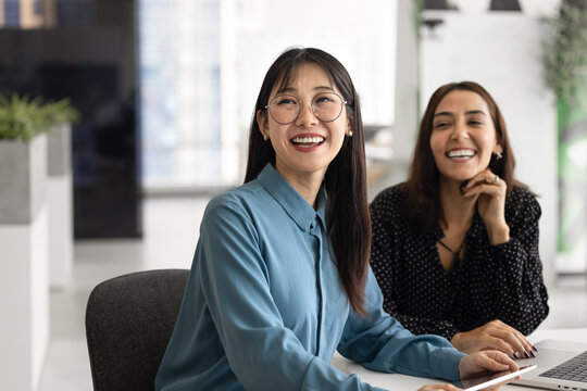 Happy young Asian business professional woman in glasses sitting at office table, working with positive Middle Eastern female coworker together, looking away, smiling, laughing