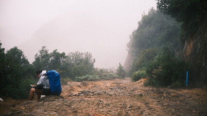 A hiker sits on a rocky mountain trail carrying a large backpack during foggy weather while checking a mobile phone in a remote outdoor landscape. © salmon