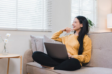 Happy young asian female sitting on comfortable sofa using laptop computer in home living room and...