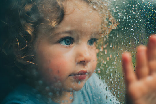 A curious child touching raindrops on a glass window, watching water droplets slide down, blurred rainy background