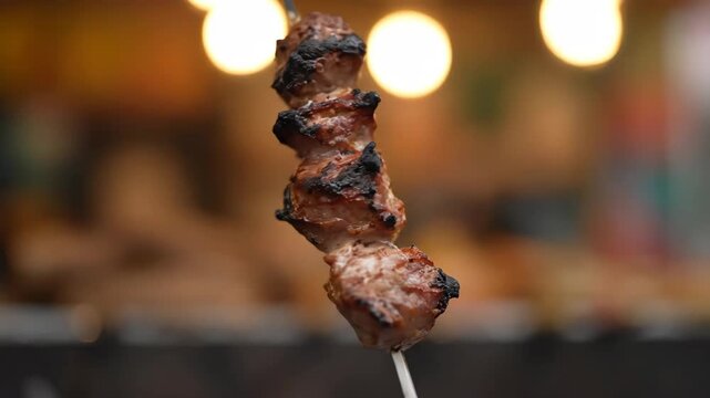 Macro shot of savory grilled meat pieces on a skewer, cooked to perfection, with a warm, blurred bokeh background suggesting a night market or street food stall environment.