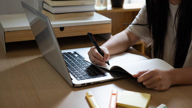 Remote worker writing notes during virtual meeting on laptop, Student studying from home taking online course notes, Close up of hands working from home office desk with pen and paper