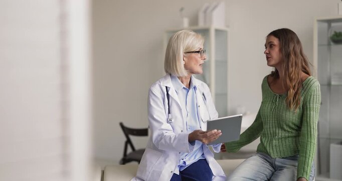 Female doctor showing health information on tablet to woman patient, explains test result, clarifying new telemedicine application use during medical consultation in clinic. Digital healthcare service