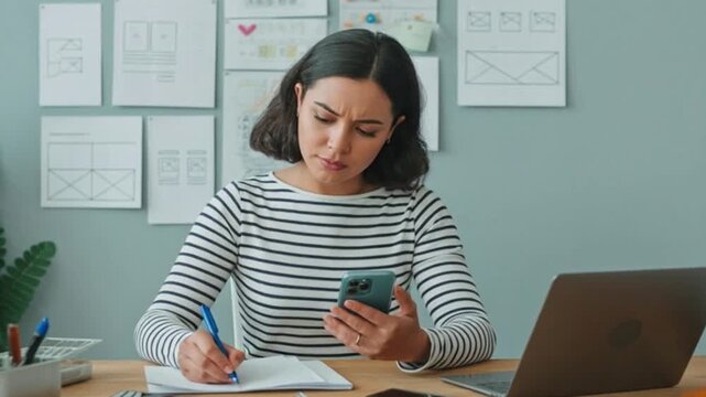 Medium vertical shot of busy young Hispanic female user interface designer in stripy shirt working on smartphone, writing notes on paper while developing wireframes and mockups  4K Video.