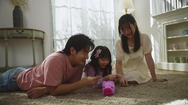 Asian Family teaching child to save money at home, parents and daughter putting coins into piggy bank. financial education, family bonding, parenting, savings, and smart money habits from childhood.