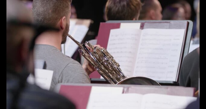 A slow motion video of a musician playing the French horn from sheet music on a music stand during a wind ensemble rehearsal