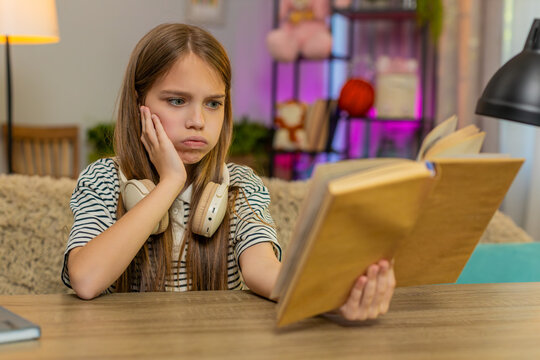 Young child girl sitting at table on home sofa looks sadly into school textbook struggling to understand the task. School kid holds head in confusion showing frustration and difficulty during homework