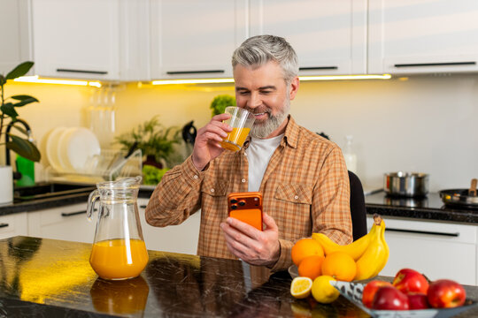 Middle-aged man pours juice into glass checks smartphone news at home kitchen during morning routine feeling happy. Mature guy drinks vitamin juice for healthy diet wellness indoors using smartphone.