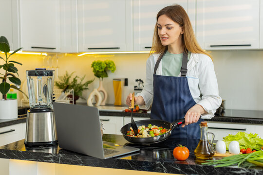 Middle-aged woman cooks fresh vegetables by easy video recipe using laptop for healthy diet meal in home kitchen. Young adult girl feels happy satisfied and focused on clean eating nutrition indoors.