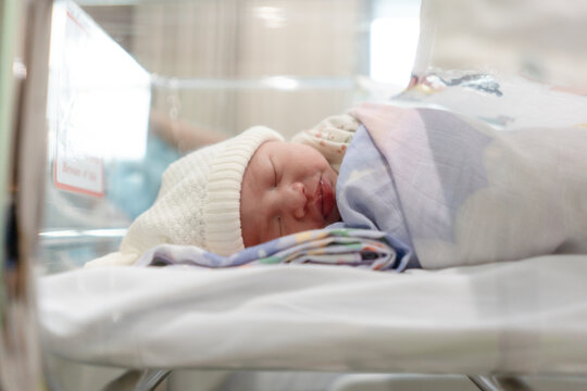 Adorable newborn baby sleeping peacefully in a hospital bassinet, wrapped in a floral swaddle blanket and wearing a white knit hat. A serene close-up shot of infancy in a bright nursery clinic.