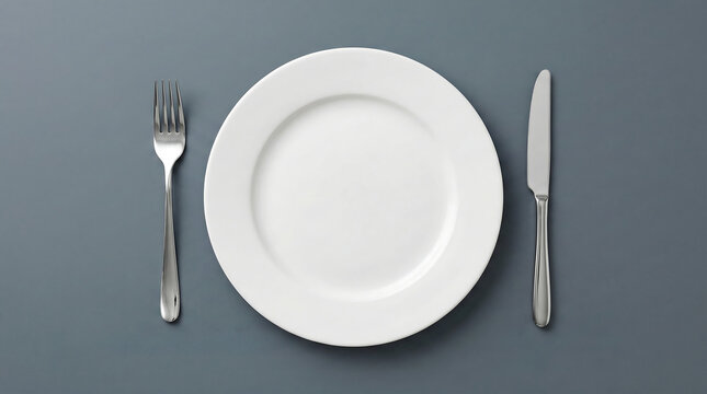 Top view of a white ceramic plate with a silver fork and knife placed on a dark grey background for a minimalist dining table concept tonight