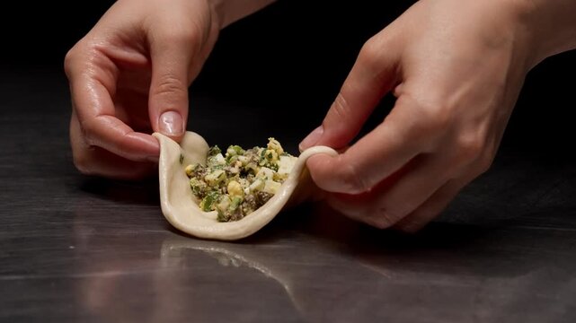 Closeup sequence of skilled hands folding and sealing a small dough parcel filled with savory vegetable stuffing on a dark reflective kitchen surface