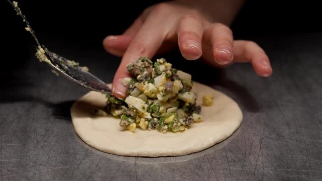 Closeup of raw dough circle on dark surface gradually filled with savory chopped vegetable and egg stuffing, highlighting homemade cooking preparation process