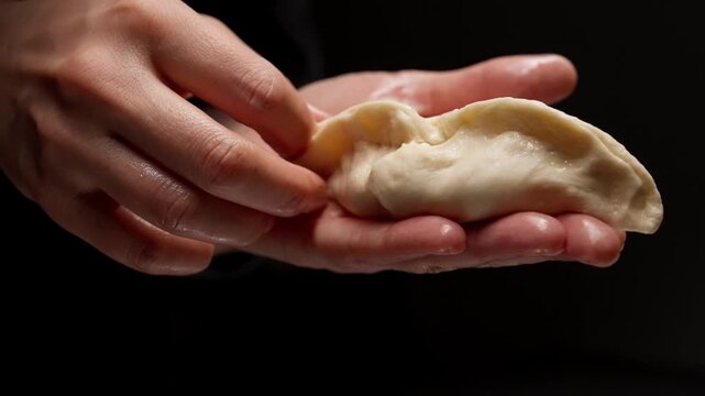 Close-up hands delicately shaping and sealing soft dough dumpling in low light kitchen, showing traditional handmade cooking process and careful finger craftsmanship