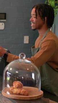 Vertical video: Smiling man in apron taking order at counter, tapping POS and adjusting pastry dome