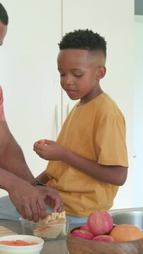 Vertical video: Slicing bread dad packing container while son in mustard nibbling snack at counter