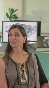 Indian woman is turning to off-camera speaker in office, smiling and discussing floorplan on screen