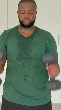 Vertical video: Picking dumbbells, man in green tee and shorts at home curling for workout, pausing