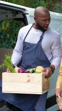 Vertical video: Sitting on van step unloading crate, man in navy apron checking produce with basket
