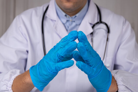 Un m&eacute;decin en blouse blanche et st&eacute;thoscope, avec des gants en nitrile bleu se tenant les mains.