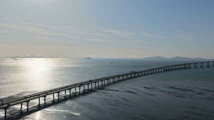 Fototapeta premium Aerial drone photography of the long curved path of Incheon Bridge stretching over the shimmering sea under a soft blue sky at golden hour in South Korea.