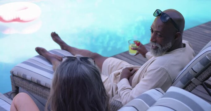 Senior couple lounging poolside, woman pointing at pink float, African American man offering toast