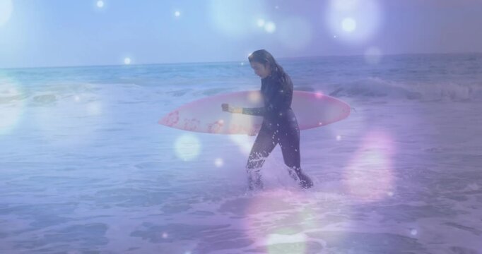 Woman surfer wading into surf, stepping deeper, adjusting pink board, preparing surfing under bokeh