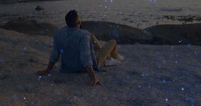 Man sitting on rock shore, camera framing starting blue VFX particles drifting around him for tech