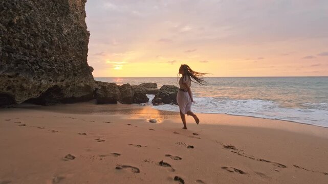 Joyful young woman with long braided dreadlocks runs barefoot across golden sand toward turquoise ocean on tropical beach at sunset. Female wears a flowing light beige maxi skirt and cropped top that