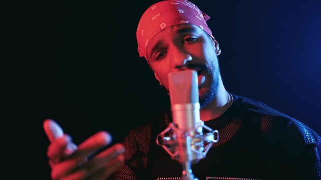 Young male artist singing and gesturing passionately into a professional studio microphone. Creative hispanic musician performing on stage with red and blue lights