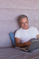 Senior man reclining against mauve headboard on bed, using laptop and wearing reading glasses