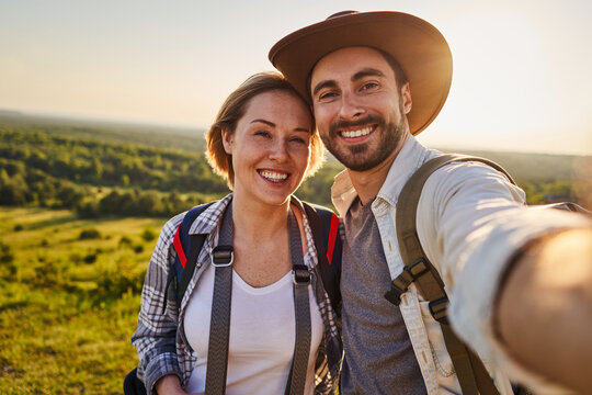 Happy couple takes selfie while hiking outdoors at golden hour with scenic countryside landscape behind them