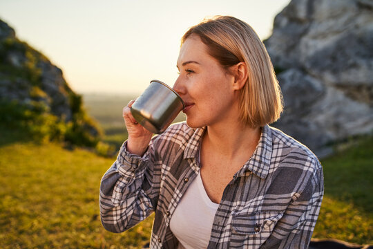 Young Woman Drinking Hot Beverage From Metal Cup While Relaxing Outdoors in Mountains at Golden Hour