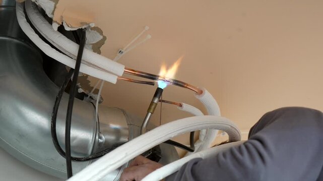 Professional hvac technician brazing copper pipes of an air conditioner unit, using a gas torch to solder the refrigerant lines during an installation or maintenance service at a construction site