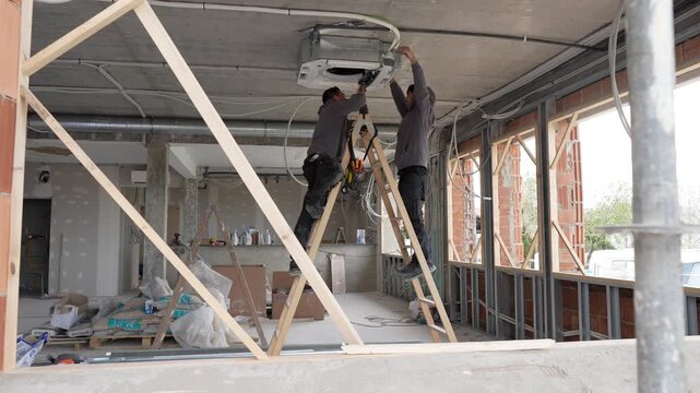 Two professional hvac technicians standing on ladders and installing a ceiling mounted air conditioner unit during the renovation of a commercial building, focusing on climate control