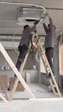 Two skilled male technicians working together on a construction site, standing on ladders to install the electrical wiring for a ceiling mounted cassette air conditioning system