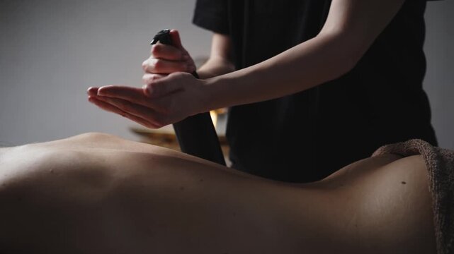 Female massage therapist applies oil from a black bottle onto the back of a client lying on a massage table in a serene indoor setting with soft lighting
