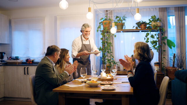 Senior man in apron serving food to his senior friends, all smiling and applauding at a cozy dinner party