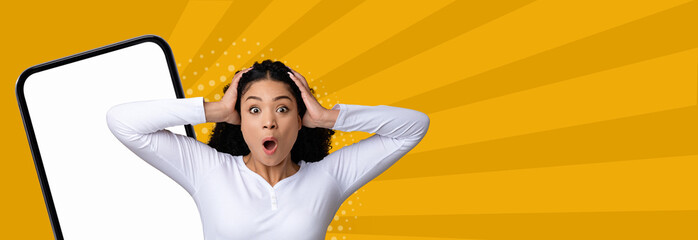 A woman expresses a look of surprise while holding her head in shock. She stands next to a large phone with a blank screen. The background features bright yellow rays. © Prostock-studio