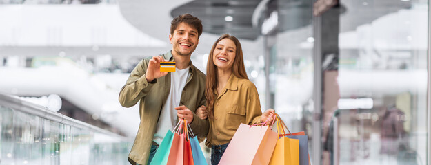 A young couple stands together inside a mall, smiling and holding shopping bags. The man shows a credit card while the woman holds colorful bags. They look happy and excited about their shopping trip. © Prostock-studio