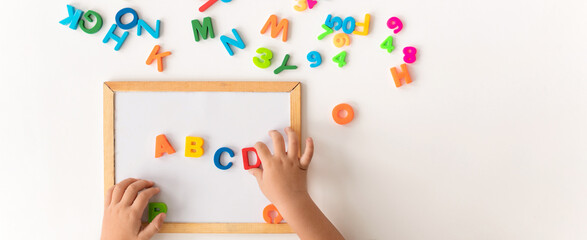 Hands of a child arrange colorful magnetic letters on a whiteboard. Some letters are scattered around on a flat surface. The activity focuses on teaching the alphabet and spelling skills. © Prostock-studio