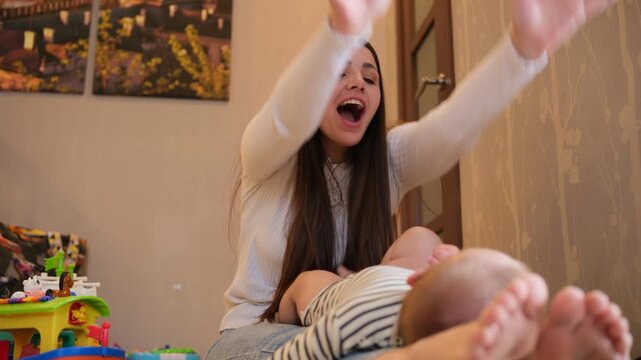 Beautiful smiling mother playing with her adorable infant son. Clapping hands and laughing together in a cozy living room. Happy family moment celebrating new life. Parenthood. And unconditional love