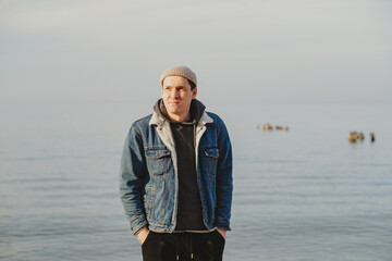 Fototapeta premium Man standing at the seaside during golden hour. Young guy wearing a denim jacket and beanie resting on the beach. Stylish traveler outdoors enjoying calm nature and peaceful atmosphere.