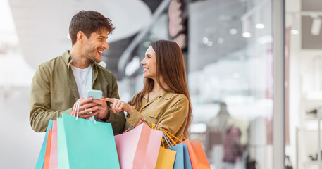 A couple stands inside a mall, smiling and talking while holding colorful shopping bags. They appear happy as they look at a phone together. It is a lively shopping day. © Prostock-studio