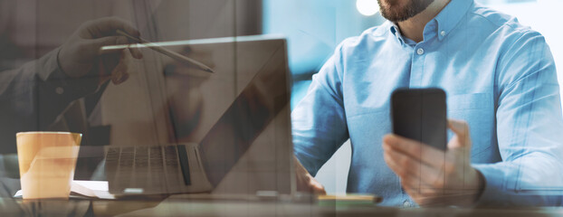 Fototapeta premium A mature male employee is engaged with his mobile phone while working on a laptop in a modern office. The setting is well-lit and has a professional atmosphere. He appears focused and productive.