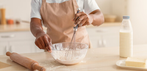 A person stands in a kitchen mixing flour and other ingredients in a bowl. There is a rolling pin and a bottle of milk on the counter. Baking preparations are in progress. © Prostock-studio